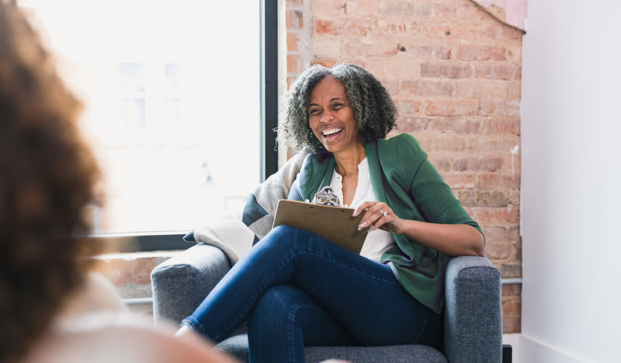 Woman holding a clipboard and laughing while talking with a patient.